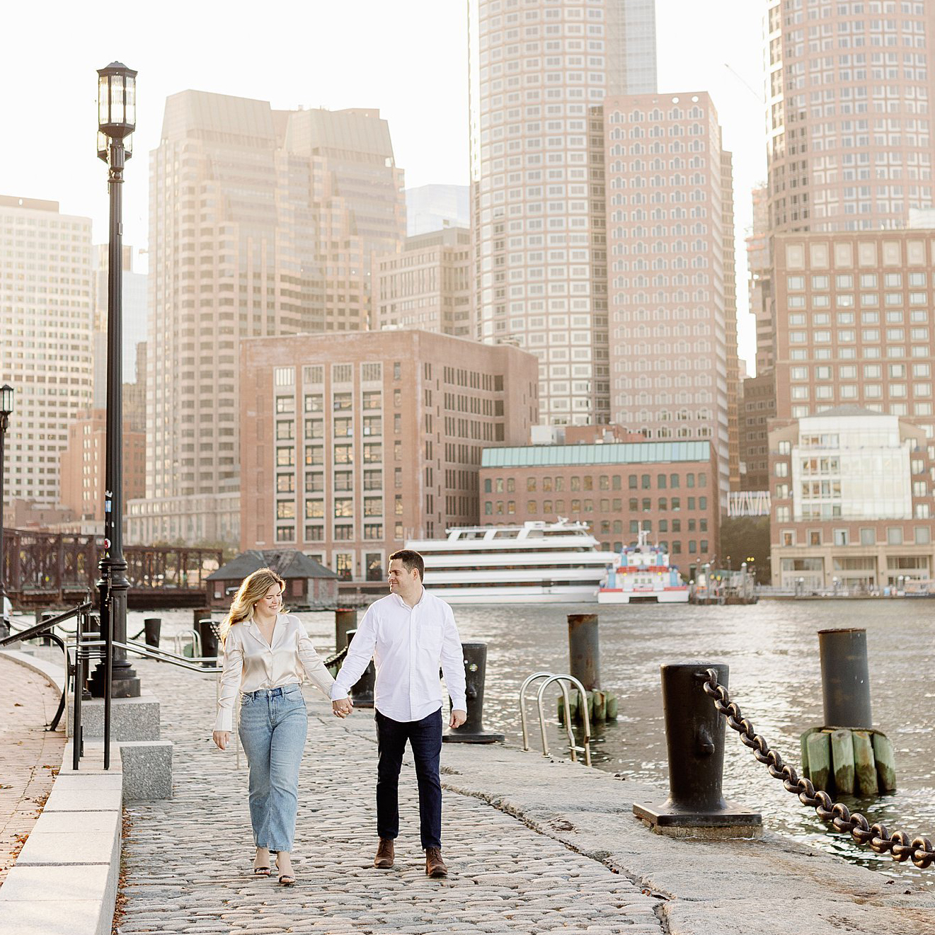 Couple walk together with the Boston skyline behind them at dusk. Deborah Zoe Photography.