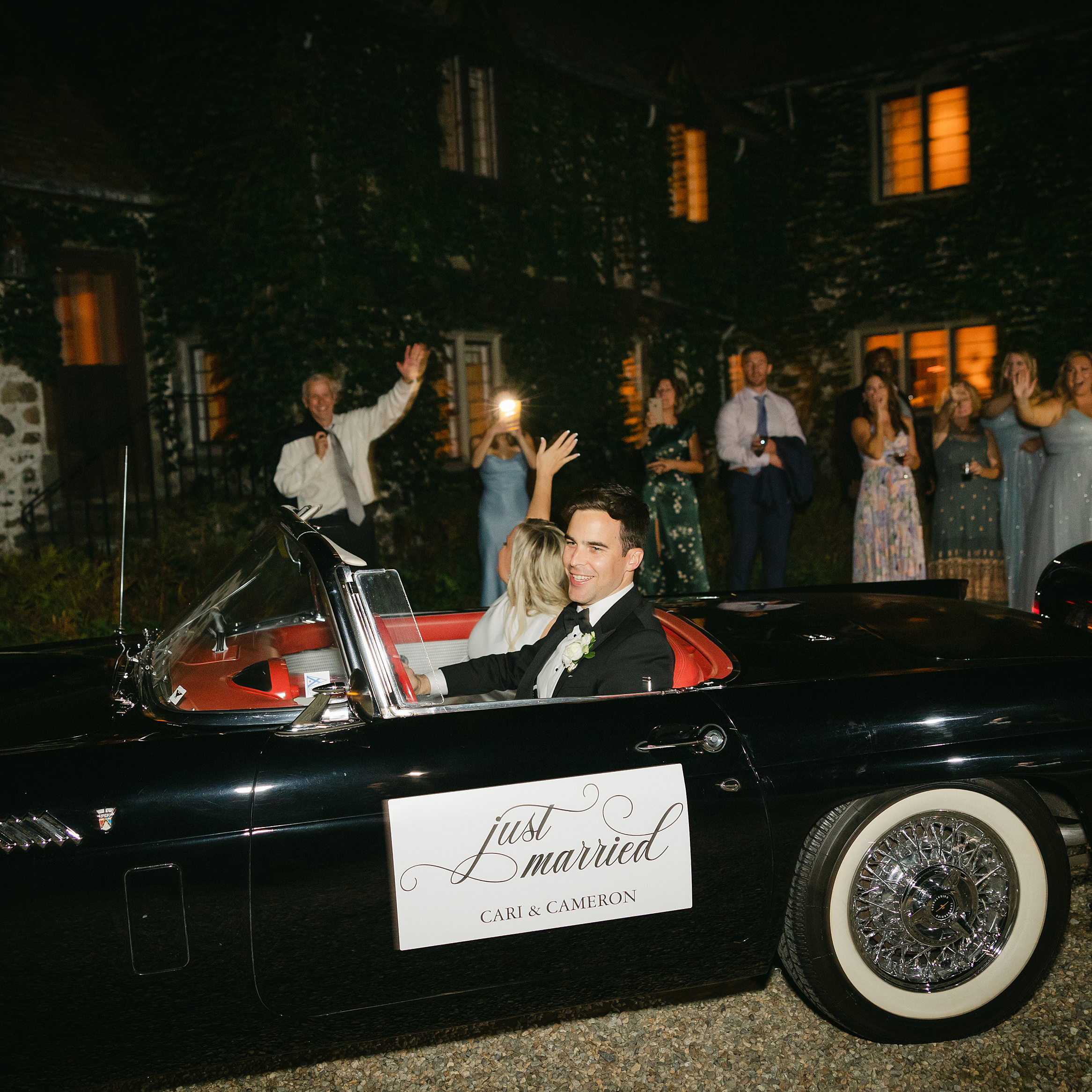 Couple leave their wedding reception in a vintage car as guests wave in the background. Deborah Zoe Photography Private Estate Wedding