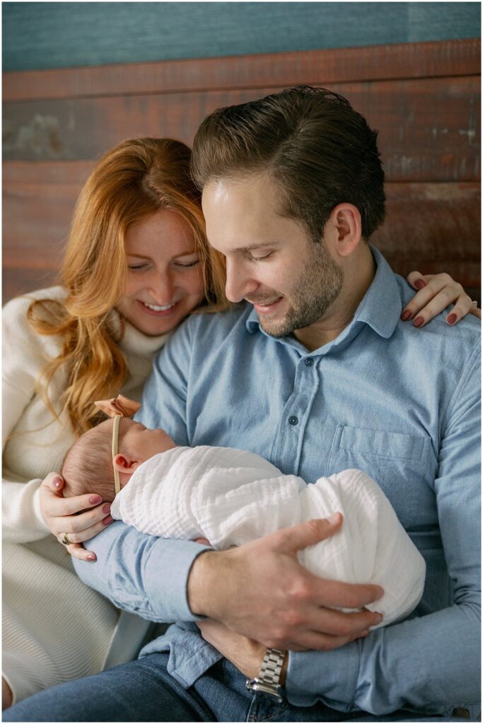 Mother and Father sit on bed and hold swaddled newborn. Boston Newborn Session Deborah Zoe Photography.