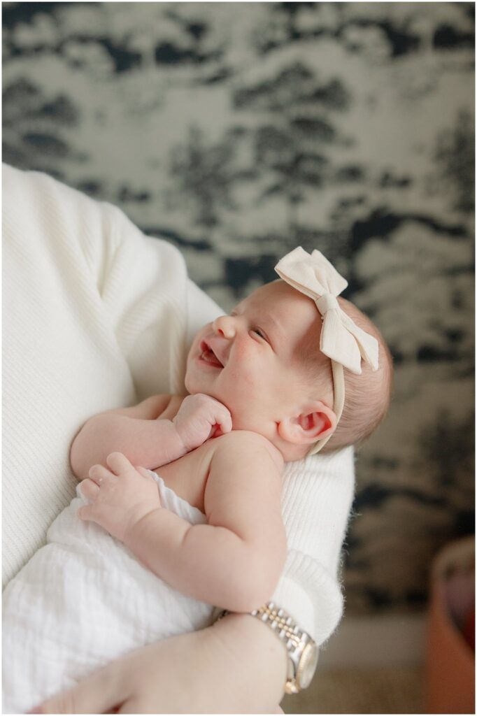 Swaddled baby smiles as wearing a bow as she is held by her Mom. Boston Newborn Session Deborah Zoe Photography