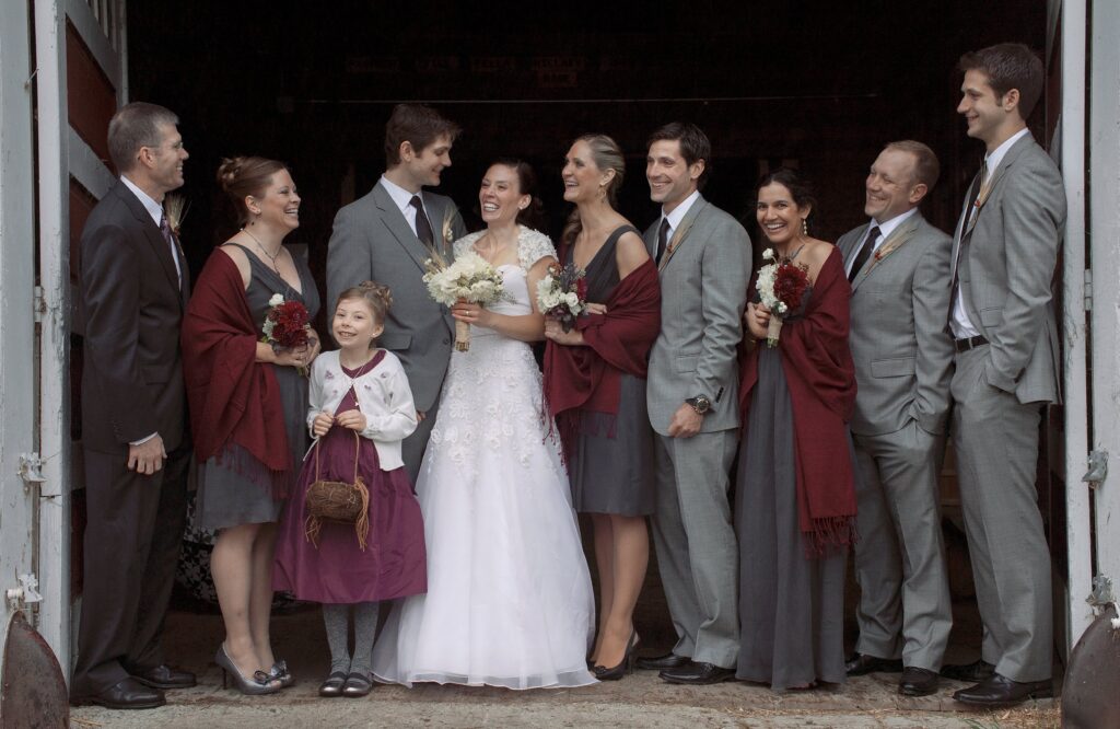 Wedding party dressed in grey and burgundy laugh together with Bride and Groom. Vermont Wedding Photographer Deborah Zoe Photography