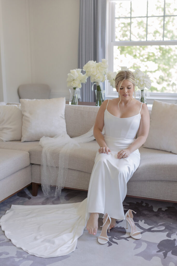 Bride sits on a couch and puts on her white heels. Modern Summer Wedding at the Newbury Boston Hotel photographed by Deborah Zoe Photography. 