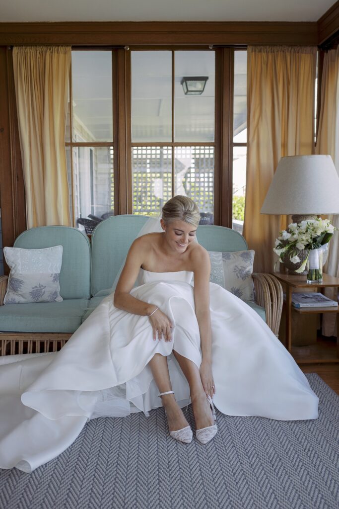 Bride seated puts on her shoes in he wedding dress. Coastal New England Wedding.