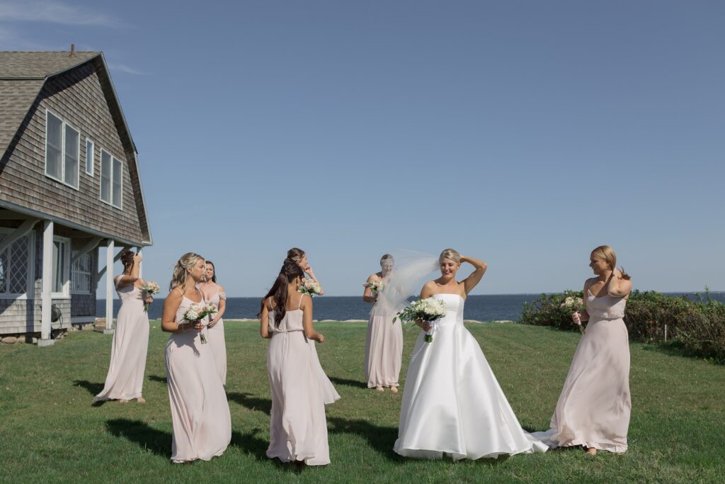 Bride holds her veil as the wind blows surrounded by her Bridesmaids. Coastal New England Wedding.