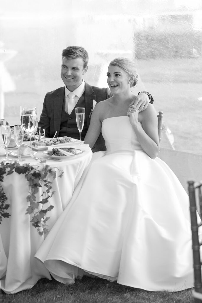 Bride and groom seated next to each other smiling during wedding toasts. Coastal New England Wedding.